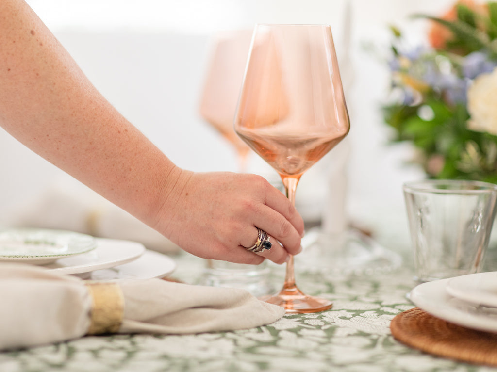 Ryan Harper Designs - Hand Placing Rose Colored Wine Glass Down On Green and White Table Setting